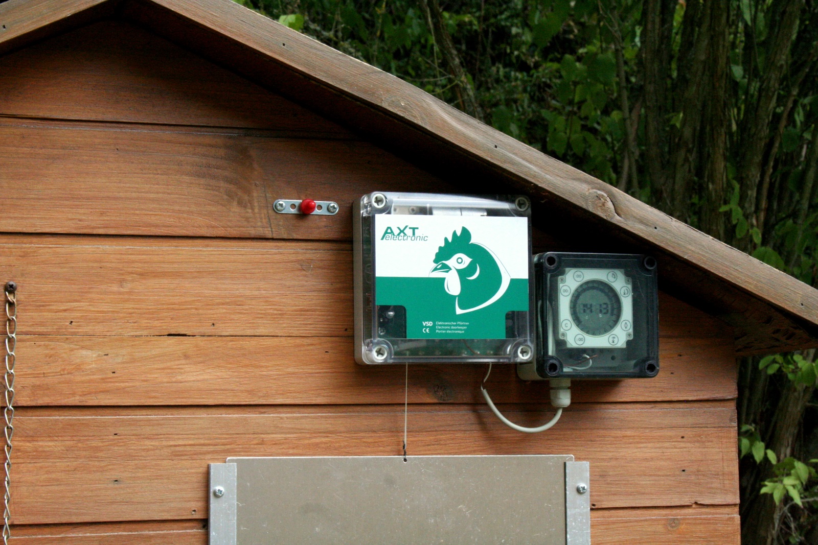 Elektronischer Pförtner VSD mit AXT electronic Hühner-Logo und externe digitale Zeitschaltuhr an brauner Holzwand eines Geflügelstalls. Uhrzeit „14:13“. Links roter Knopf zur manuellen Steuerung, darunter teils silberner Schieber.
