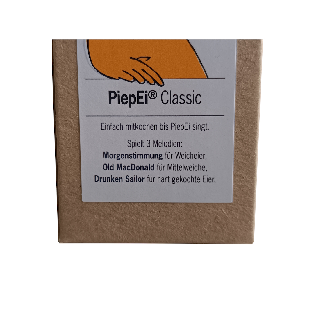 Verpackung des PiepEi Classic. Das Etikett auf der braunen Box beschreibt die musikalische Eieruhr, die beim Kochen singt. Die drei Melodien 'Morgenstimmung', 'Old MacDonald' und 'Drunken Sailor' signalisieren weiche, mittelweiche und hart gekochte Eier.