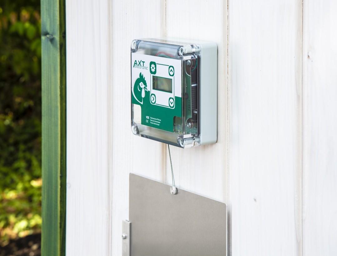 Ein AXT electronic Hühnerklappenöffner mit grünem Logo und Display ist an einer weißen Holzfassade montiert. Darunter hängt eine metallene Schiebetür, die an einem Seil befestigt ist. Im Hintergrund sind unscharfe grüne Pflanzen zu sehen.