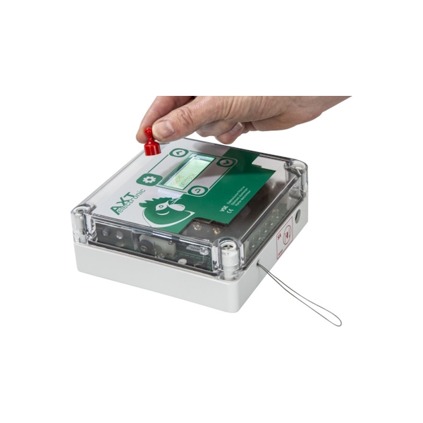 Eine Hand drückt einen roten Knopf auf einem weißen Gerät mit transparenter Abdeckung. Innen ist eine grüne Platine "AXT electronic" mit Hühnerlogo. Ein kleines Display zeigt "Germany". Rechts ragt eine Drahtschlaufe heraus.