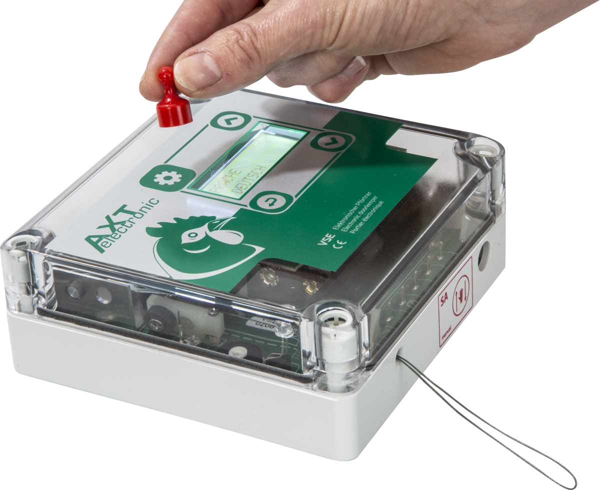 Weißes, durchsichtiges Steuergerät für automatische Hühnerklappe. Eine Hand hält einen kleinen roten Magneten zur Bedienung darüber. Display: 'SPRACHE DEUTSCH'. Grünes Label: 'AXT electronic' mit Hühnerlogo. Dünner Draht seitlich.