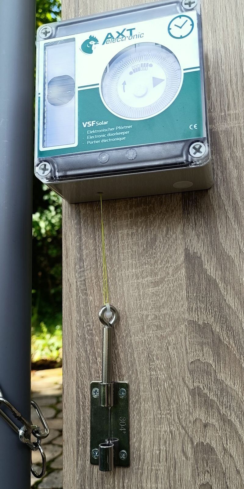 Detailansicht eines automatischen Weidetoröffners BZ3 an einer Holzwand. Oben die AXT electronic Steuerung mit Solarpanel und mechanischer Zeitschaltuhr. Grüner Seilzug zum Edelstahl-Bolzen zum Verriegeln/Öffnen.