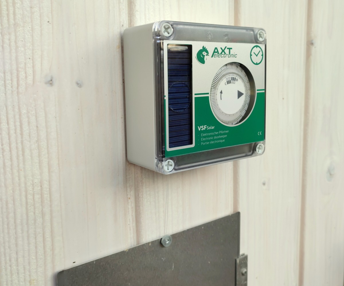 Elektronischer Hühnerstalltüröffner von AXT electronic, Modell VSF Solar, an heller Holzwand. Mit blauem Solarpanel und rundem Zeitschaltuhr-Zifferblatt mit Zahlen und Pfeil im transparenten Gehäuse. Unten hängt ein Faden zur Metalltür.