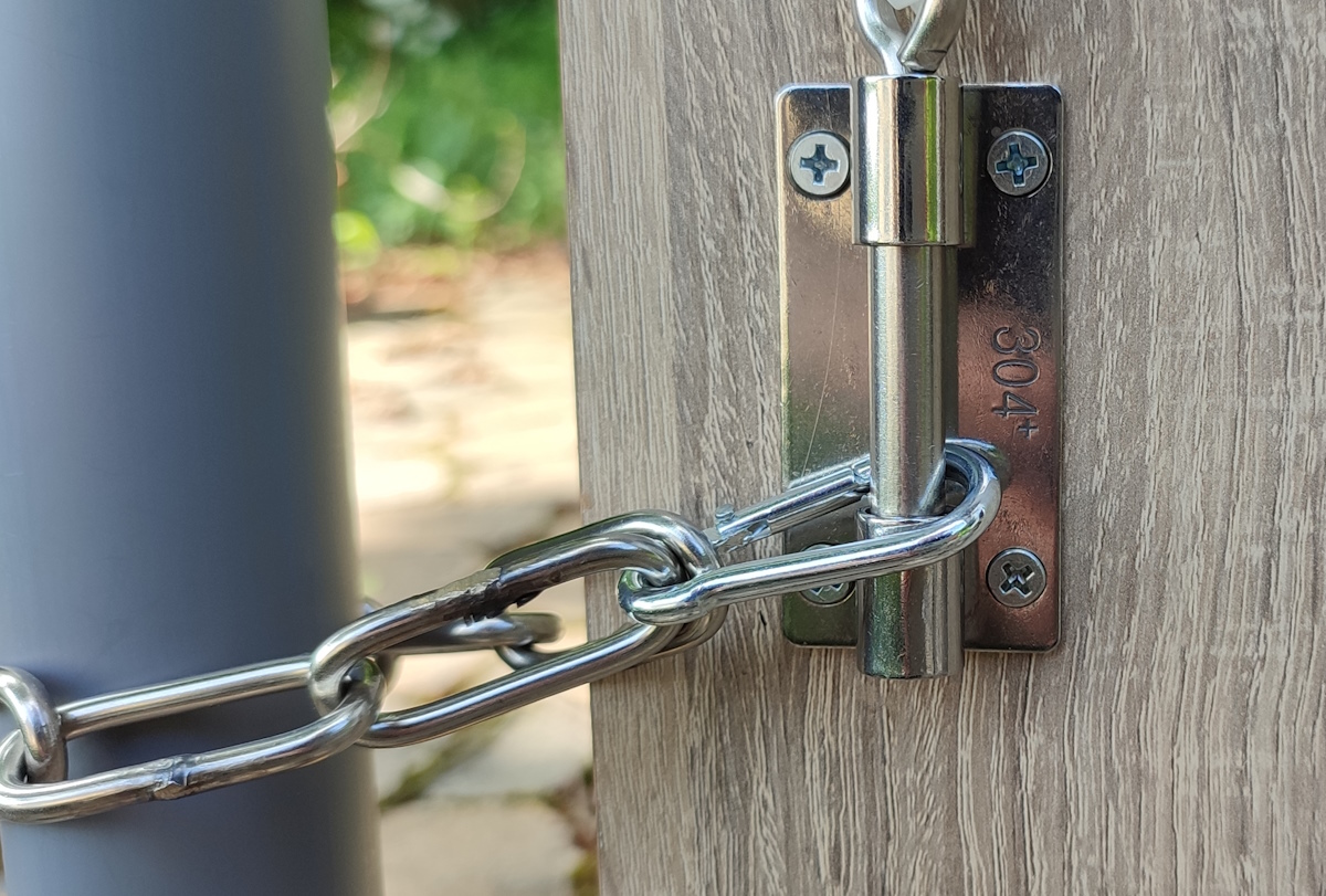 Nahaufnahme eines glänzenden Edelstahlbolzens in robuster Metallführung auf heller, holzähnlicher Oberfläche. Eine massive silberne Kette umschlingt den Bolzen und verschließt ein Weidetor. „304+“ ist eingraviert.