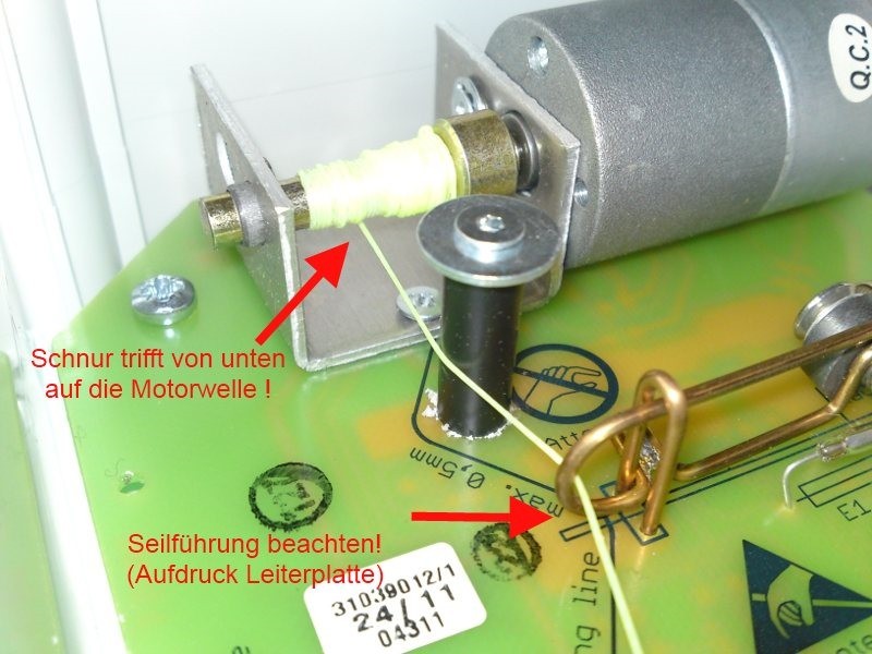 Detailansicht einer grünen Leiterplatte mit Elektromotor. Eine dünne hellgelbe Schnur ist um die Motorwelle gewickelt und kommt von unten. Sie folgt einer aufgedruckten Linie als Führung. Rote Pfeile und Text markieren die korrekte Montage.
