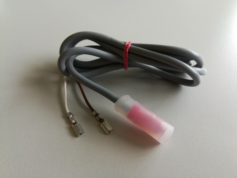 Aufgerolltes graues Verlängerungskabel mit rotem Gummiband. An einem Ende zwei dünne Kabel (weiß, braun) mit blanken Metallsteckern. Am anderen Ende transparente Kappe mit rosa Bauteil. Externes SA-Verlängerungskabel.