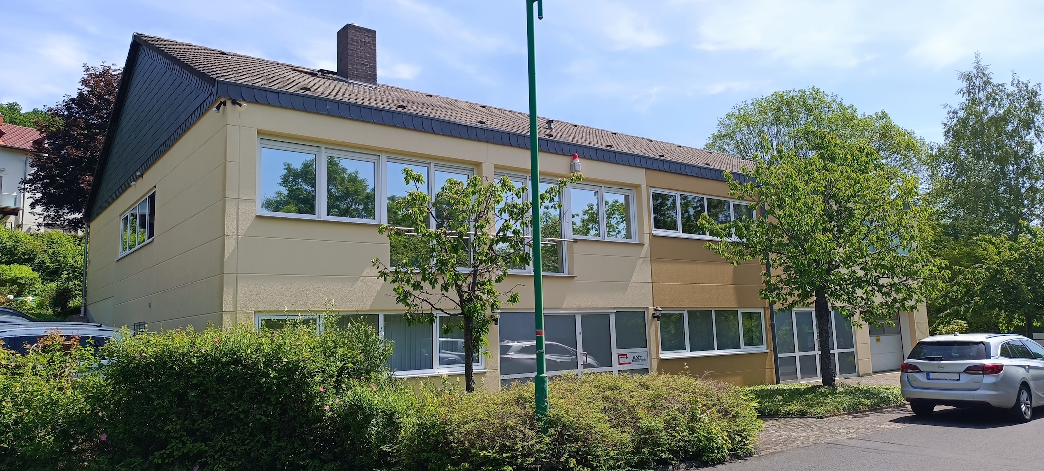 Modernes, zweistöckiges Gebäude mit hellbeiger Fassade und vielen Fenstern, die grüne Bäume und blauen Himmel spiegeln. Dichte Büsche und kleinere Bäume vorne. Rechts parkt ein silberner Kombi auf gepflastertem Hof.