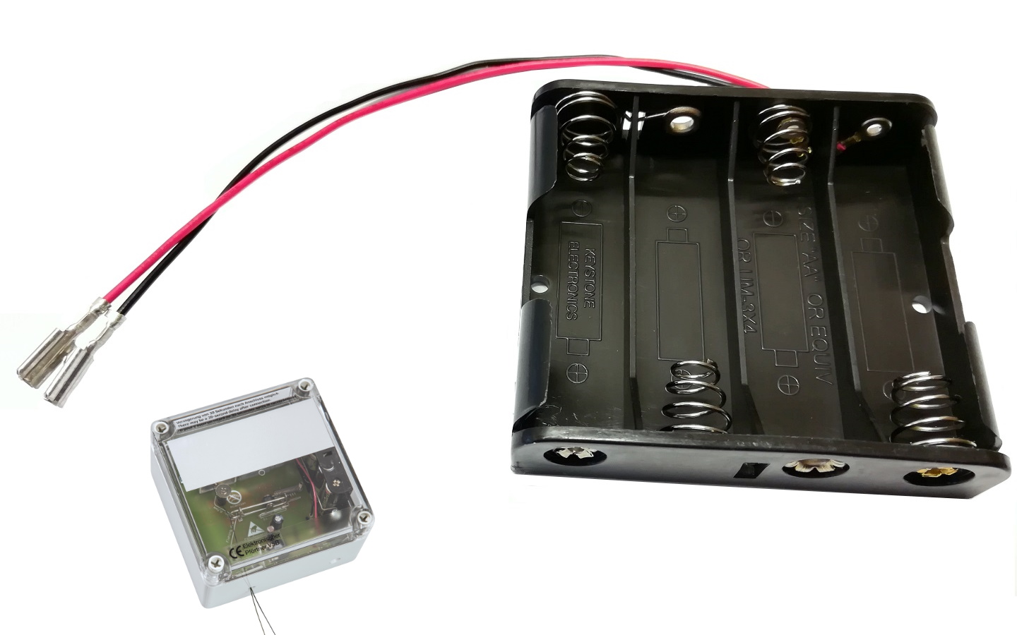 Schwarzer Batteriehalter für vier AA-Batterien mit silbernen Federkontakten im Vordergrund. Zwei rote und schwarze Kabel mit Flachsteckern ragen heraus. Links ein transparentes Elektronikgehäuse (Pfrtner VSBb) als Ersatzteil.