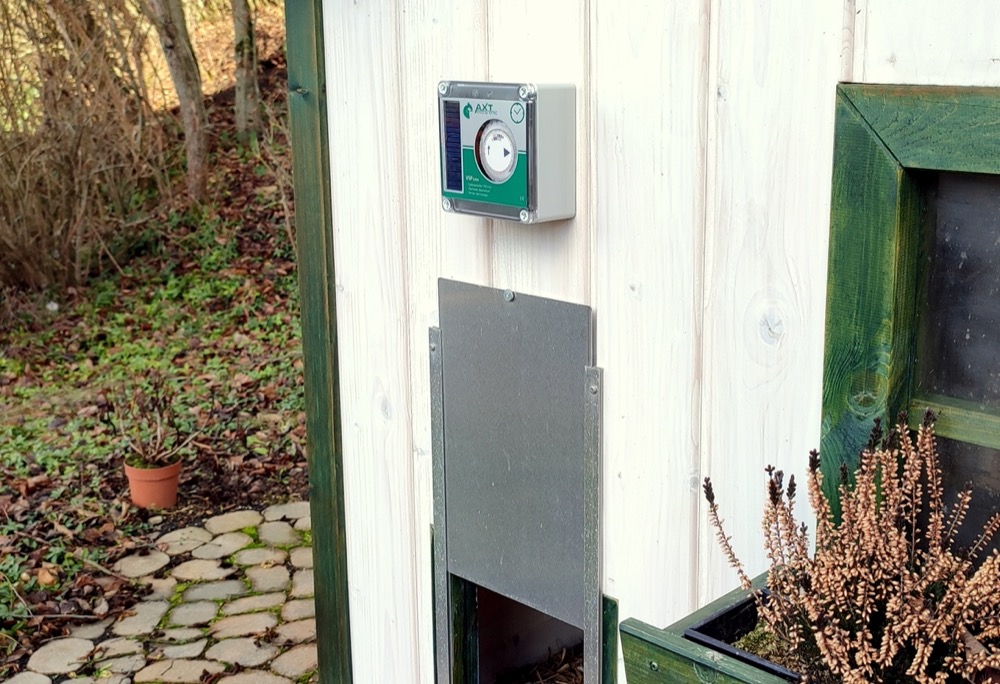 AXT VSF Hühnerklappensteuerung mit Zeitschaltuhr und Solarpanel an weißer Stallwand; darunter halb geöffnete Schiebetür. Grüner Fensterrahmen und Natur am Rand.
