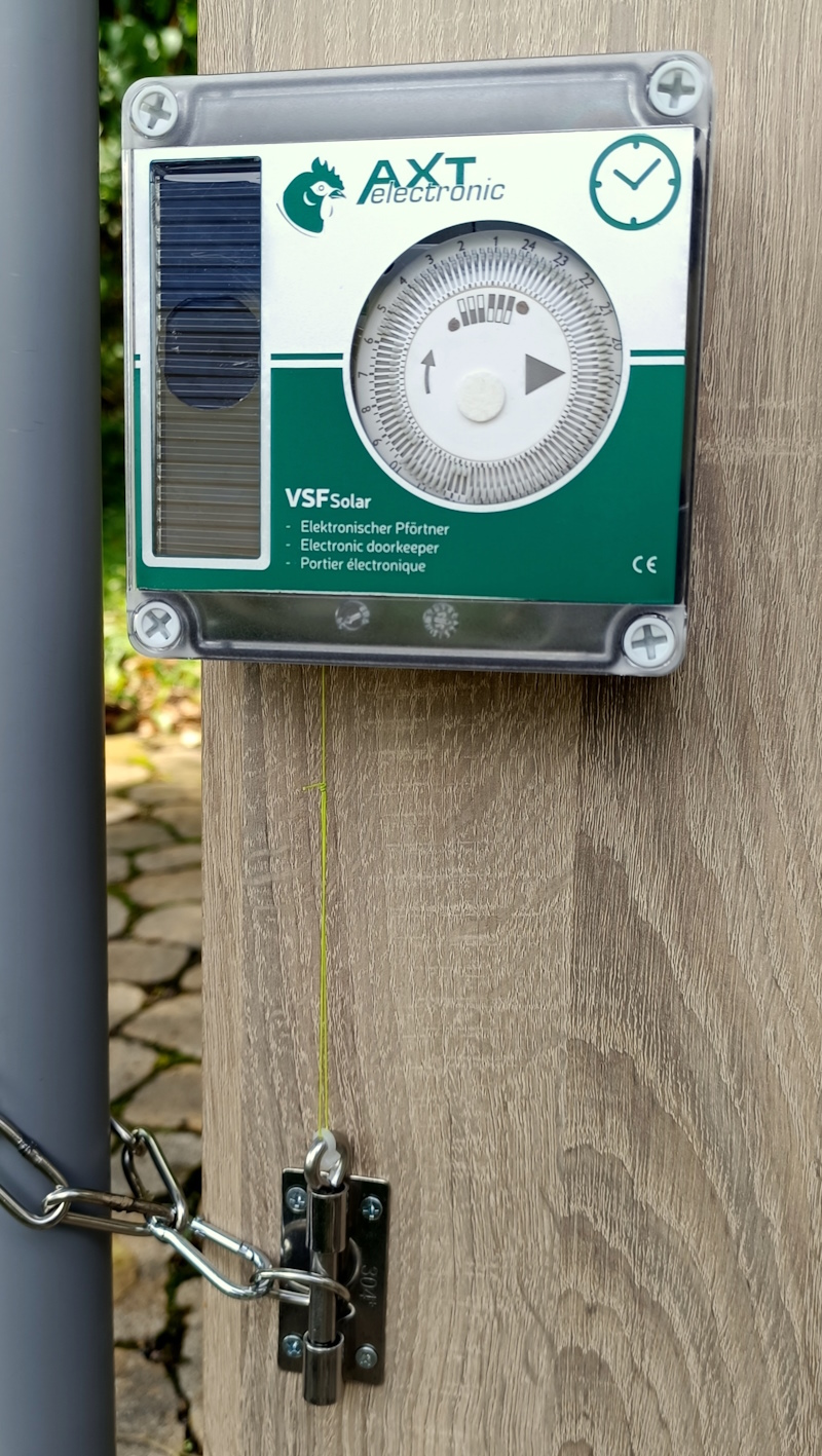 An einer Holzwand montierte AXT electronic VSF Solar-Steuerung für Weidetore mit analoger Zeitschaltuhr und Solarpanel. Ein gelber Seilzug verbindet sie mit einem 8mm Edelstahlbolzen, der eine Kette zum Schließen sichert.
