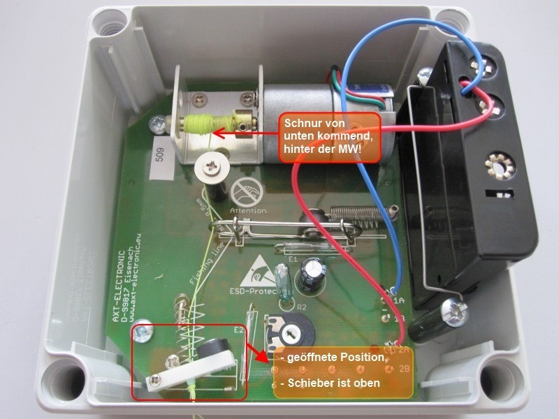 Nahaufnahme einer weißen Elektronikbox mit offenem Deckel: grüne Platine mit Bauteilen, roten und blauen Kabeln sowie kleinem Motor mit gelb-grüner Schnur. Markierungen zeigen Schnurführung hinter dem Motor und offenen weißen Schieber mit Feder.